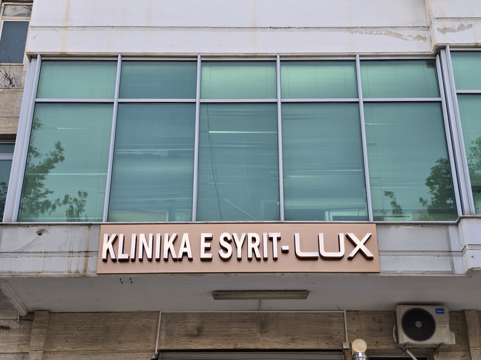Copper-toned 3D letters reading KLINIKA E SYRIT-LUX on a beige panel below a green-glass facade