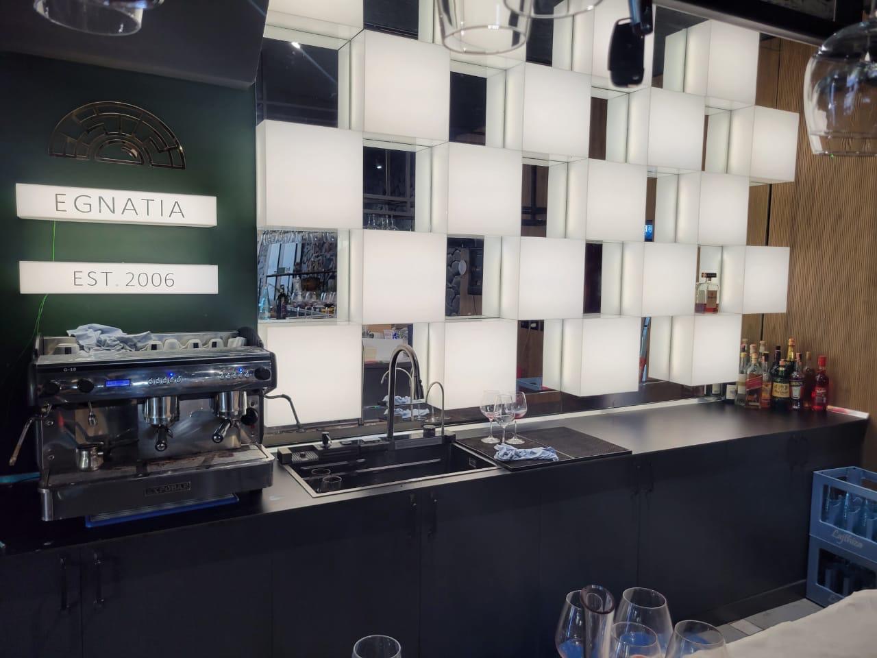 Wall of illuminated white cubes behind the counter with EGNATIA EST. 2006 panels on a green wall