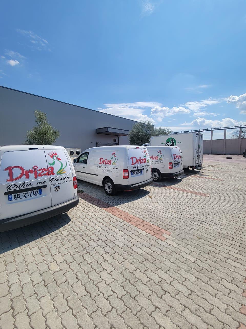 Three white Driza vans with the rooster logo and the Rritur me Pasion slogan, lined up outside the plant