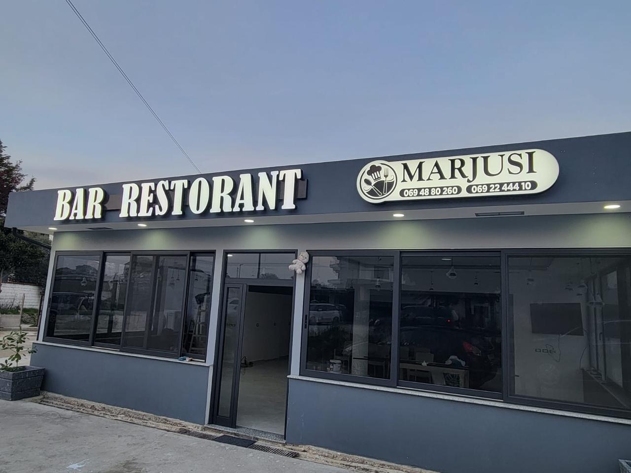 Illuminated BAR RESTORANT letters and an oval Marjusi lightbox with phone numbers, lit at dusk